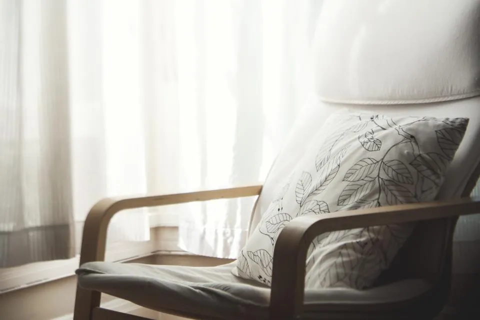 Armchair with a patterned cushion beside a sheer-curtained window in a rented room